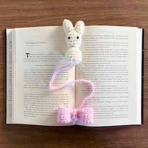 Könnte beinhalten: Ein weißes, gehäkeltes Hasen-Lesezeichen mit einer rosa Schleife. Das Lesezeichen liegt auf einem geöffneten Buch.