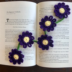 May include: A purple crocheted flower bookmark with a yellow center, attached to a green stem. The bookmark is placed on an open book with text about a circus.