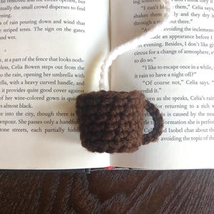 Puede incluir: Un marcap&aacute;ginas de taza de caf&eacute; de ganchillo marr&oacute;n con un lazo de hilo blanco. El marcap&aacute;ginas est&aacute; sobre un libro abierto con texto sobre una noche lluviosa.