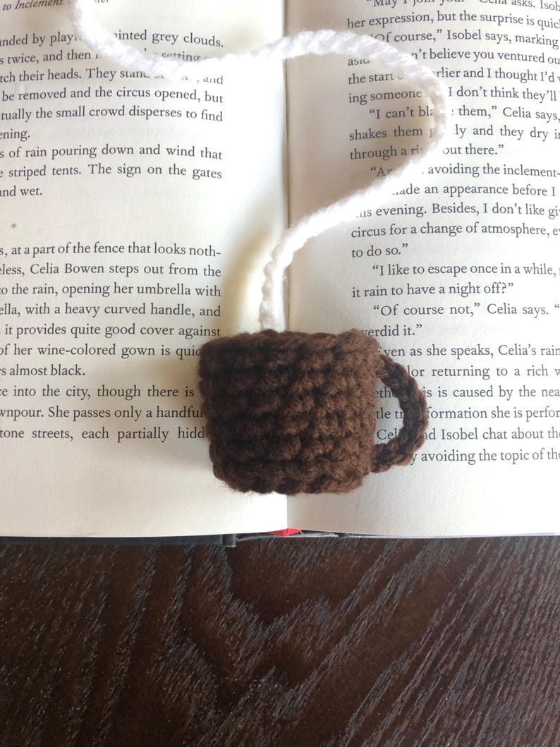 Puede incluir: Un marcap&aacute;ginas de taza de caf&eacute; de ganchillo marr&oacute;n con un lazo de hilo blanco. El marcap&aacute;ginas est&aacute; sobre un libro abierto con texto sobre una noche lluviosa.