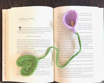 May include: A close-up of a book with a crocheted bookmark in the shape of a green heart and a purple calla lily. The book is open to a page with the title "The Magician's Umbrella" and the text "Closed Due to Inclement Weather".