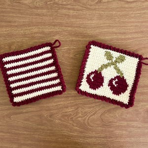 May include: Two crocheted pot holders, one with white and burgundy stripes and the other with a burgundy cherry design on a white background.