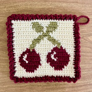 Puede incluir: Un agarrador de olla tejido a crochet con un borde rojo y un diseño de dos cerezas con tallos y hojas verdes.
