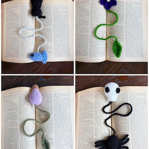 May include: Four handmade crochet bookmarks in an open book. The bookmarks feature a black cat with a blue fish, a purple flower with a green leaf, a lavender bulb with a green leaf, and a white skull with a black spider.