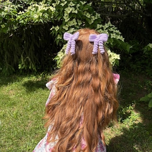 May include: A person with long, wavy auburn hair styled with two lavender crochet hair bows. The bows are in a bow shape and are attached to the hair. The person is wearing a floral dress and is in a garden setting.