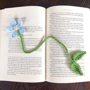 Peut inclure: Un marque-page en crochet bleu en forme de fleur avec une tige et des feuilles vertes. Le marque-page est placé sur un livre ouvert avec du texte sur un cirque.