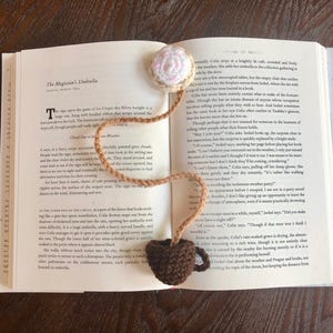 Può includere: Un segnalibro all'uncinetto con un design a forma di tazza di caffè e cupcake. Il segnalibro è marrone e beige ed è attaccato a un libro con il testo "The Magician's Umbrella" visibile.