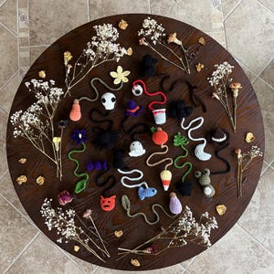 May include: A collection of handmade crochet Halloween-themed ornaments, including ghosts, pumpkins, bats, and other festive shapes. The ornaments are arranged on a dark wooden surface, surrounded by dried flowers and petals. The colors are vibrant, with orange, black, white, and purple.