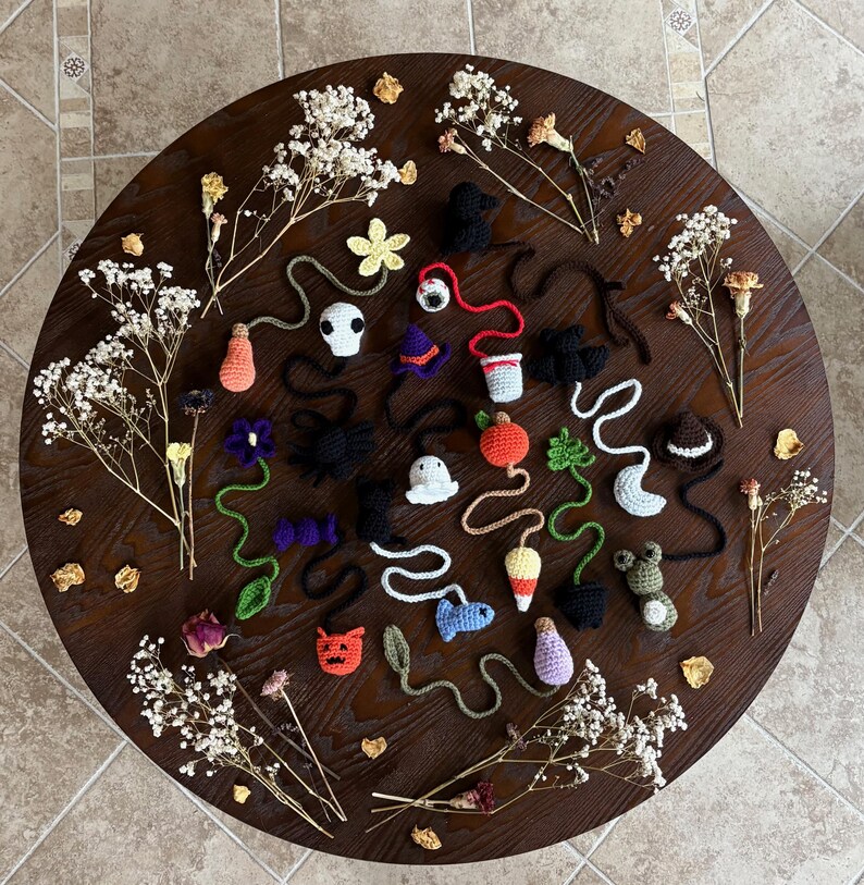 May include: A collection of handmade crochet Halloween-themed ornaments, including ghosts, pumpkins, bats, and other festive shapes. The ornaments are arranged on a dark wooden surface, surrounded by dried flowers and petals. The colors are vibrant, with orange, black, white, and purple.
