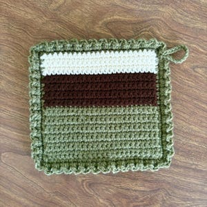 May include: A green crocheted pot holder with a white and brown striped design. The pot holder has a scalloped edge and a loop for hanging.