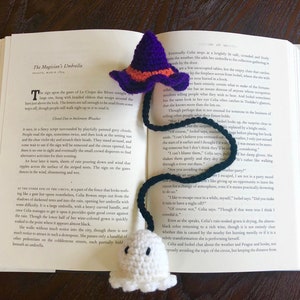 May include: A crocheted ghost and witch hat bookmark. The ghost is white and the witch hat is purple with orange trim. The bookmark is placed on an open book with the text "The Magician's Umbrella" visible.