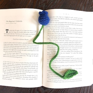May include: A blue and green crocheted bookmark shaped like a flower with a blue flower head and a green stem and leaves. The bookmark is placed on an open book with text about a circus.