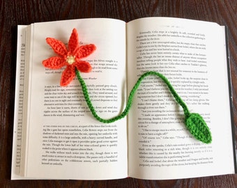 Puede incluir: Un marcapáginas de ganchillo en forma de flor con un tallo y hojas verdes. La flor es naranja y amarilla, y el tallo y las hojas son verdes. El marcapáginas está sobre un libro abierto.