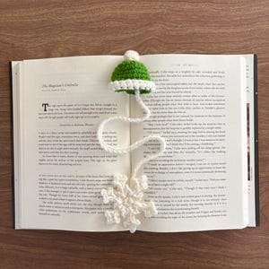 May include: A handmade crochet bookmark featuring a small green and white hat with a white tassel and a white snowflake. The bookmark is placed in an open book, adding a touch of whimsy to reading.