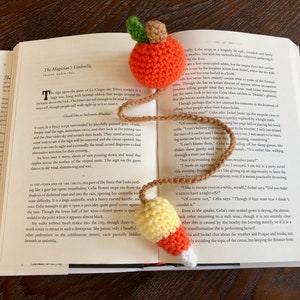 Puede incluir: Primer plano de un marcapáginas de ganchillo con un diseño de maíz amarillo y naranja en un extremo y un diseño de manzana roja y naranja en el otro extremo. El marcapáginas está sobre un libro abierto.