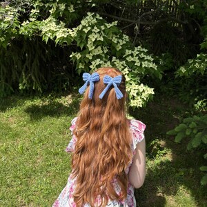 May include: Two light blue crochet hair bows are attached to long, wavy auburn hair. The person is wearing a floral dress with pink and white flowers. The background features green grass and white flowers.