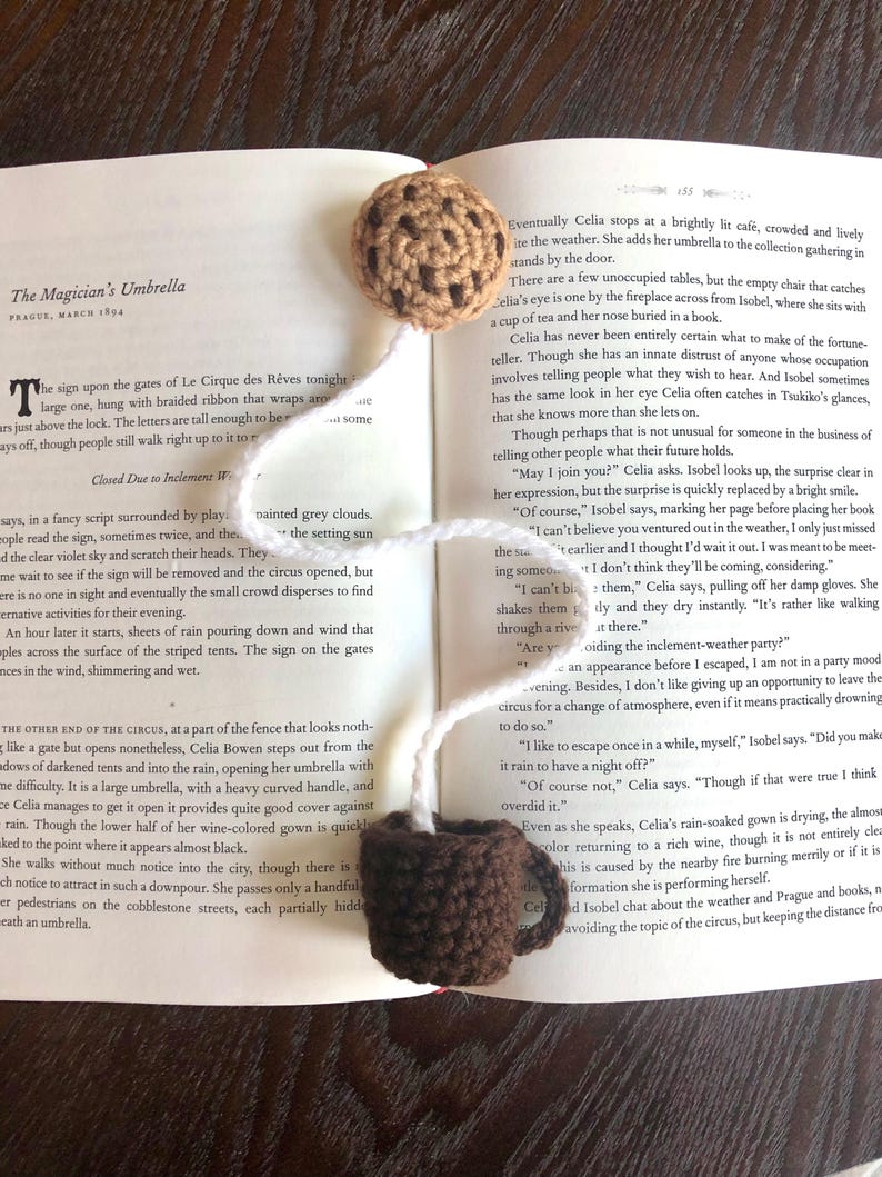 Puede incluir: Primer plano de un libro abierto con un marcap&aacute;ginas de galleta de crochet. El libro est&aacute; abierto en una p&aacute;gina con texto sobre una adivina y un circo. El texto dice "The Magician's Umbrella, Prague, March 1894".