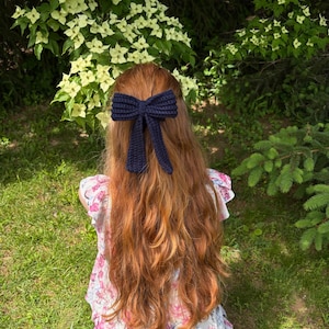 May include: A navy blue crochet bow hair accessory. The bow is attached to long, wavy auburn hair. The person is wearing a floral print dress while sitting in a grassy area with green foliage and white flowers in the background.