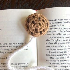 Puede incluir: Un marcap&aacute;ginas de galleta con chispas de chocolate tejido a crochet con hilo marr&oacute;n y puntadas marr&oacute;n oscuro. El marcap&aacute;ginas est&aacute; colocado en un libro abierto con texto sobre un circo.