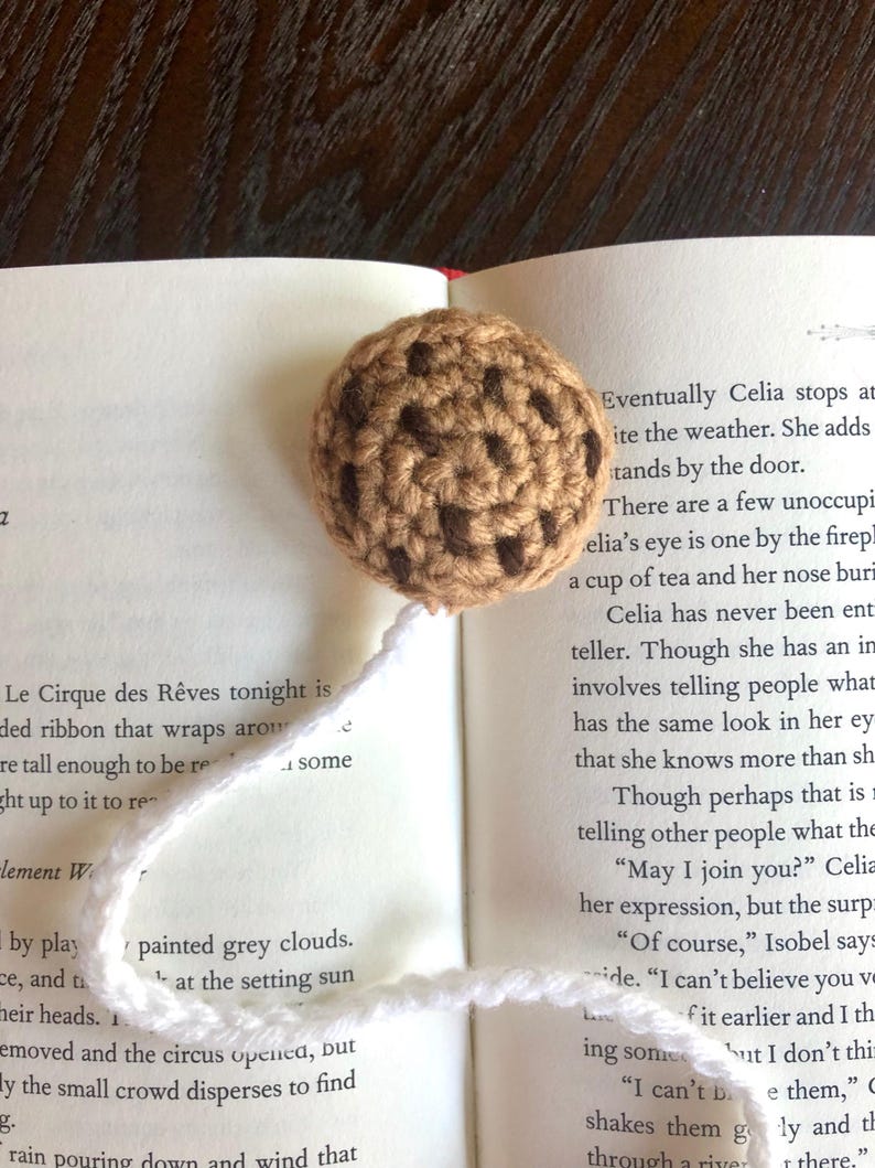 Puede incluir: Un marcap&aacute;ginas de galleta con chispas de chocolate tejido a crochet con hilo marr&oacute;n y puntadas marr&oacute;n oscuro. El marcap&aacute;ginas est&aacute; colocado en un libro abierto con texto sobre un circo.