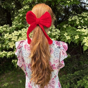 May include: A bright red, crocheted bow hair accessory. The bow is tied at the base of long, auburn hair. The person is wearing a floral print dress with pink and purple flowers.
