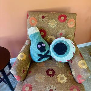 May include: Two crochet pillows rest on a floral patterned armchair. One pillow is shaped like a poison bottle, light blue with a skull and crossbones. The other is an eyeball with a black pupil and red accents.
