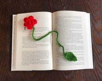 Puede incluir: Un marcapáginas de ganchillo hecho a mano con forma de rosa roja con tallo y hoja verdes, colocado en las páginas abiertas de un libro. Un accesorio único para los amantes de la lectura.