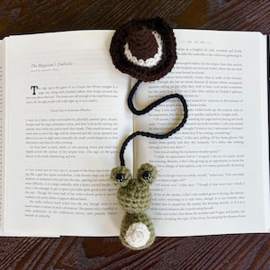 Puede incluir: Un marcapáginas de rana de ganchillo hecho a mano con un sombrero marrón y un cordón negro, colocado en un libro abierto. La rana es verde con detalles blancos y ojos grandes. El libro está abierto en una página con texto.