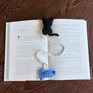 Può includere: Segnalibro all'uncinetto fatto a mano con un gatto nero in cima e un pesce blu in basso, collegati da un cordino bianco e grigio. Il segnalibro è posto su un libro aperto con testo visibile.