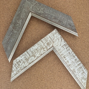 May include: Two L-shaped picture frame corner samples. The top sample features a dark grey finish with a weathered, textured surface. The bottom sample features a white finish with a distressed, wood-grain texture. Both frames have a stepped profile.
