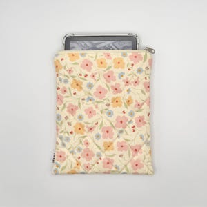 May include: A quilted cream-colored tablet sleeve with a floral pattern in pink, yellow, and blue. The sleeve has a zipper closure and a small tag with text. An e-reader is partially visible inside the sleeve.