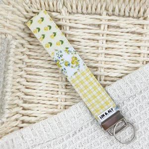 May include: A fabric key fob with a silver-tone metal clasp and key ring. The key fob features three patterned sections: yellow floral, white floral with blue accents, and yellow gingham. The text "LIN & ALY COLLECTION" is visible.
