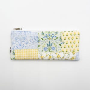 May include: A rectangular, quilted pencil case with a zipper closure. The case is made of patchwork fabric in various patterns, including floral, gingham, and damask designs in shades of blue and yellow. The text "LIN & ALY" is printed on the side.