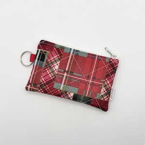 May include: A rectangular quilted pouch featuring a red, green, and white plaid pattern. The pouch includes a silver-toned metal zipper closure, a circular metal key ring attached to a red fabric tab on the left side, and a small white tag labeled LIN & ALY.