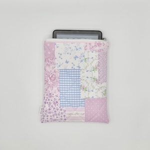 May include: A quilted tablet sleeve featuring a patchwork design of various light purple, blue, and white floral, gingham, and polka dot patterns. The rectangular fabric case has a white zipper closure at the top and a small white brand tag on the side.