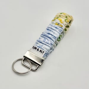 May include: A fabric wristlet key holder with a floral pattern in blue, yellow, and green. The key holder has a silver metal clasp and key ring. The text "LIN & ALY COLLECTION" is printed on a white label.