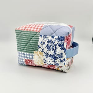 May include: A patchwork fabric cosmetic bag with a light blue handle. The bag features a variety of patterned fabrics, including gingham, floral, and striped designs. The bag has a white zipper closure and a quilted texture.