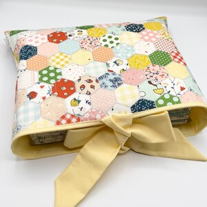 May include: A rectangular fabric pouch featuring a patchwork design of multicolored hexagons with various patterns including strawberries, florals, and gingham. The pouch has a pale yellow fabric trim and a matching pale yellow fabric bow tie.