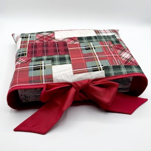 May include: A rectangular fabric cover featuring a patchwork of red, green, and white plaid patterns. The cover has a solid red fabric border along the opening and a large, tied red fabric bow centered at the front edge of the opening.
