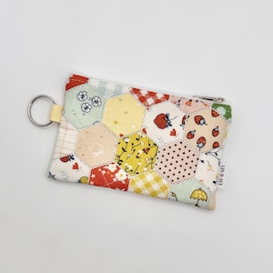 May include: A rectangular fabric zipper pouch featuring a quilted hexagonal patchwork design. The pattern includes various prints such as strawberries, yellow gingham, floral motifs, and polka dots. A silver metal keyring is attached to the side.