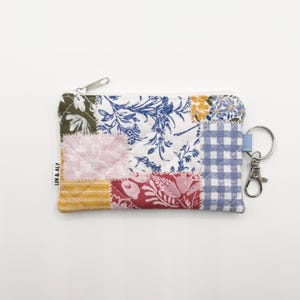 May include: A patchwork coin purse with a zipper closure and a key ring. The purse features a variety of floral and patterned fabrics in shades of blue, pink, red, yellow, and green. The text "LIN & ALY" is printed on the side.