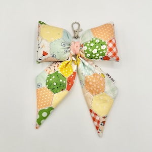 May include: A fabric bow keychain featuring a patchwork design of hexagonal patterns in various colors including yellow, green, orange, and white. The bow includes floral, polka dot, and gingham prints with a silver metal lobster clasp and a small tag.
