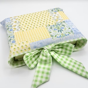 May include: A quilted book cover with a patchwork design in yellow, blue, and white floral and gingham patterns. A green and white gingham bow secures the cover. The cover is designed to protect a book.