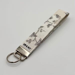 May include: A fabric key fob with a floral pattern in gray and white. The key fob has a silver metal clasp and key ring. The text "LIN & ALY COLLECTION" is printed on a small white tag.