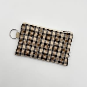 May include: A rectangular quilted zipper pouch featuring a brown, black, and cream plaid pattern. The pouch includes a silver-toned metal zipper closure and a circular metal key ring attached to the side with a small fabric loop. A white tag with the text LIN & ALY is sewn into the side seam.