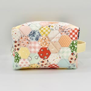 May include: A quilted pouch featuring a patchwork design of hexagonal fabric pieces in various pastel colors and patterns, including strawberries, gingham, polka dots, and floral prints, with a solid light yellow side handle and a zipper closure.
