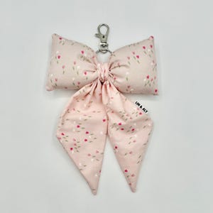 May include: A light pink fabric bow keychain featuring a repeating pattern of small red and white tulips. The bow is attached to a silver-colored metal lobster clasp and includes a small white fabric tag with the black text LIN & ALY.
