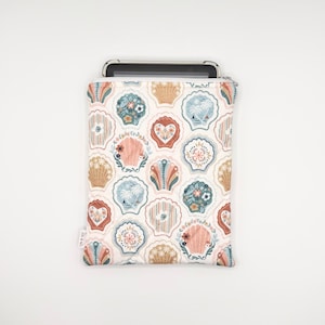 May include: A rectangular fabric sleeve with a white background featuring a repeating pattern of stylized seashells in shades of peach, teal, and tan. The shells contain various floral and palm tree motifs. A black tablet is partially inserted inside.