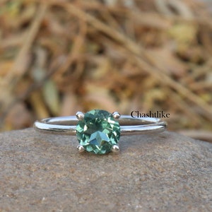 Può includere: Un anello d'argento con una pietra ovale verde. L'anello è posto su una roccia grigia.