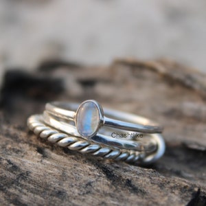 May include: A set of three silver rings featuring a moonstone gemstone. The rings are stacked on top of each other, with the moonstone ring at the top. The rings are displayed on a wooden surface.